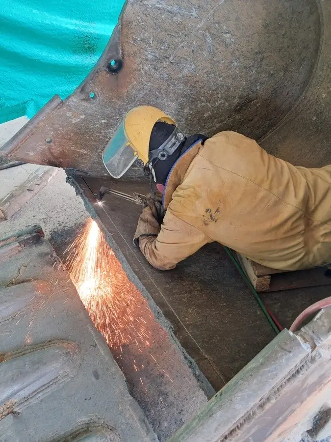 Técnico de SIMA ejecutando oxicorte con careta y polainas de cuero dentro de un balde de excavadora